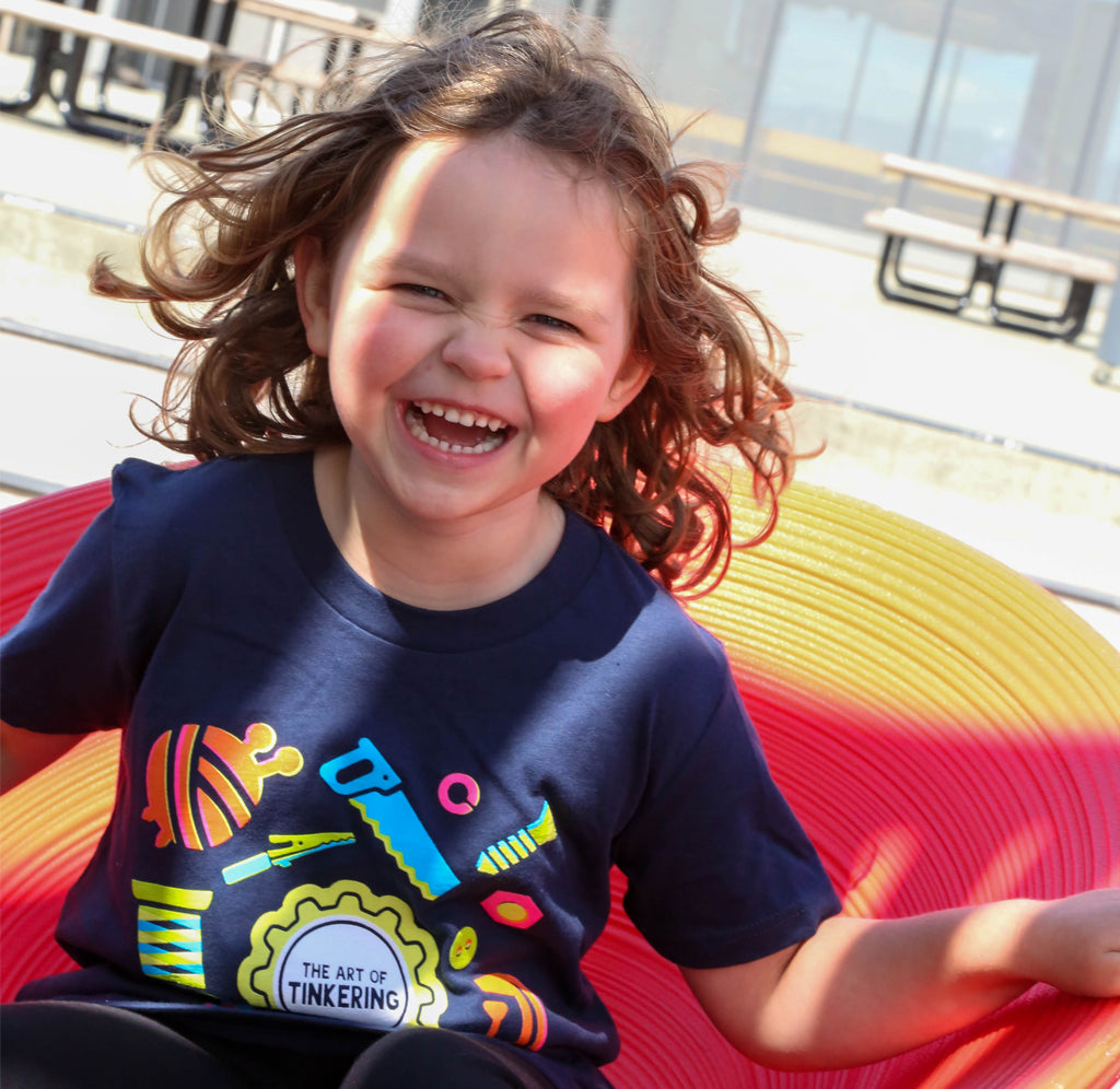 A young child is sitting on a red chair; she is wearing a blue shirt with neon tools in green, teal, pink, orange, and yellow on the front surrounding the gear-shaped Art of Tinkering logo in yellow, white, and black. 