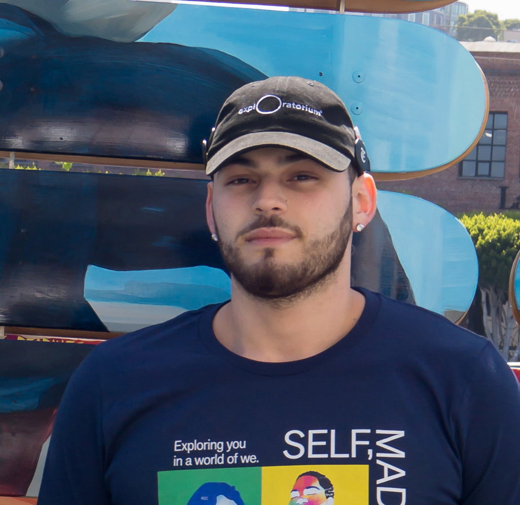 Male in front of the Exploratorium's skateboard exhibit in a black baseball cap with Exploratorium across the front in white.
