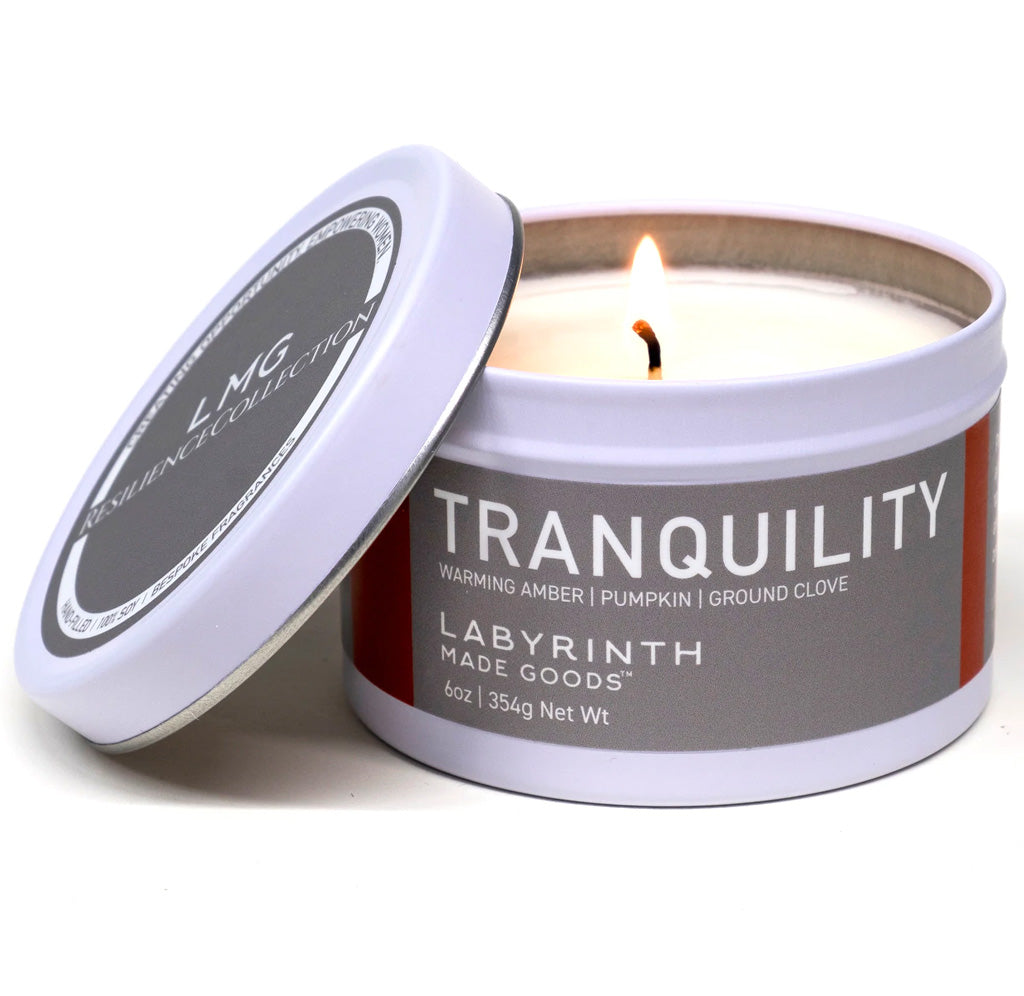 The tin for the candle is white with a silver label with white text that reads Tranquility warming amber, pumpkin, and grand clove; the white candle is lit.