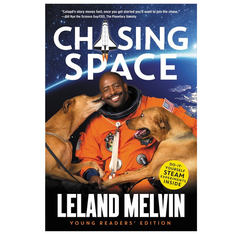 Leland Melvin is pictured in front of an image of earth from space in his orange space suit with two dogs on his lap; one is licking him.