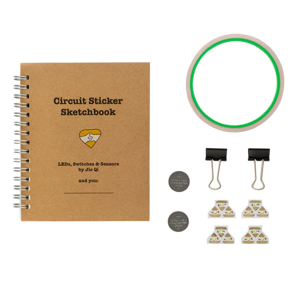 The kit contents are laid out side by side. The circuit sticker sketchbook cover is natural cardboard color; two silver coin cell batteries, a roll of copper tape, two black binder clips, and four sets of three triangular-shaped circuit stickers lie next to the sketchbook.