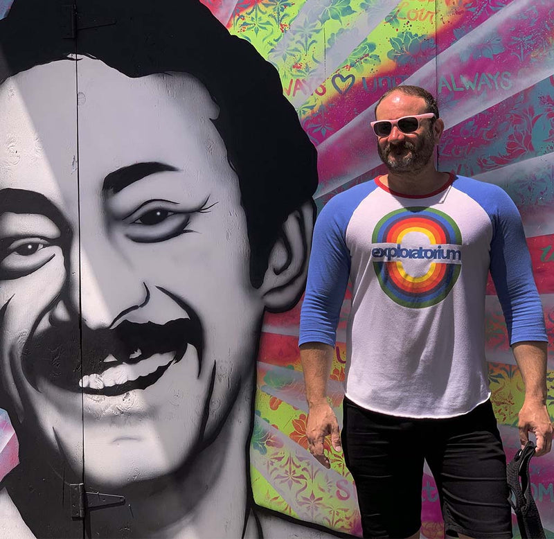 A male is standing next to a mural of Harvey Milk in San Francisco's Castro district. He is wearing a white long sleeve jersey with blue sleeves and a red collar with a  circular rainbow design. Exploratorium is underneath in blue. 