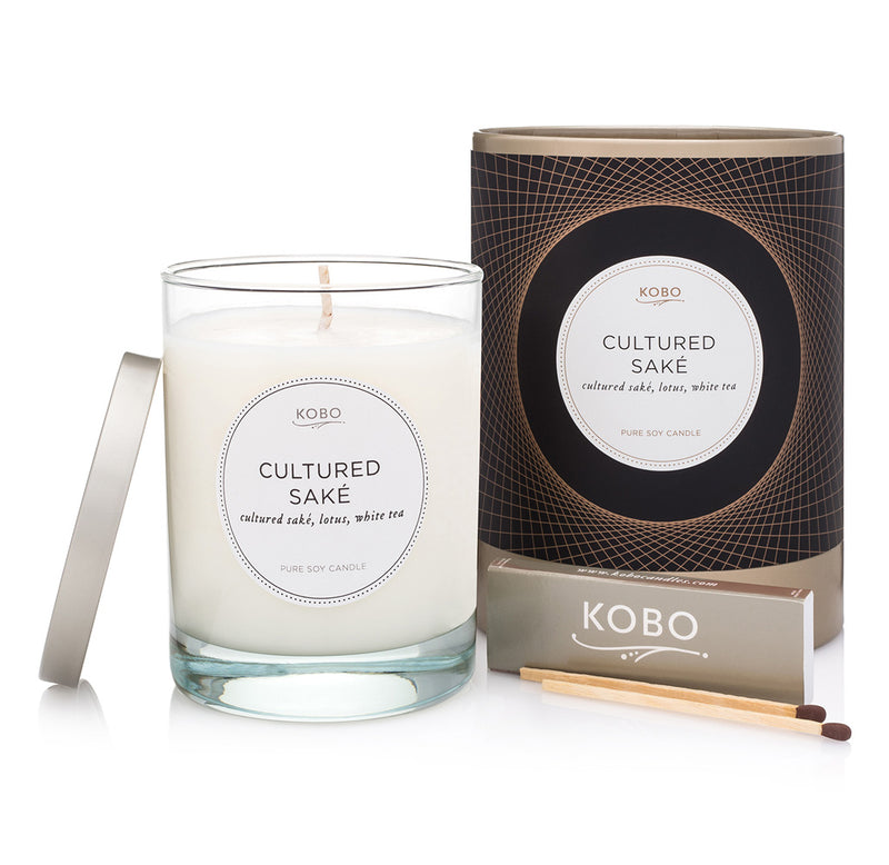 The white Cultured Saké candle sits in a glass container with a silver lid; the packaging is cylindrical with gold, black, and white colors. A small set of silver matches sits in front.