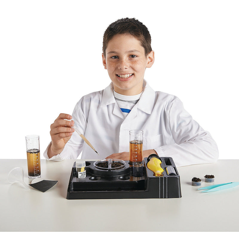 A young man in a lab coat is holding a dropper over dirt to test it for threats in the soil. There is a tray with graduated cylinders of dirty water, a microscope, and other lab equipment.
