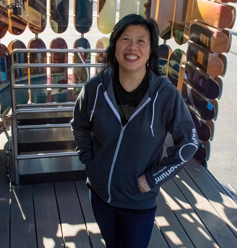 Female wearing a gray hoodie with Exploratorium in white down the left sleeve.