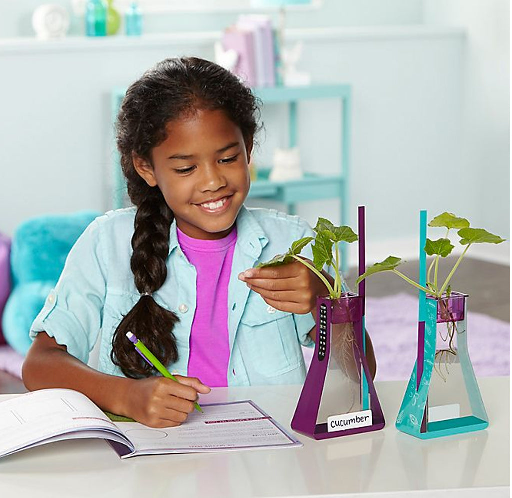 A young girl is sitting at a table examing the two plants that she has grown in the water in the blue and purple water flasks.