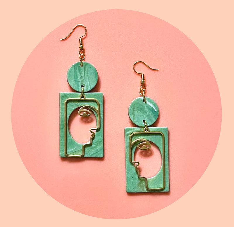 The earrings are green circles with a rectangle below with profile faces made out of wire in a cut-out in the middle. 