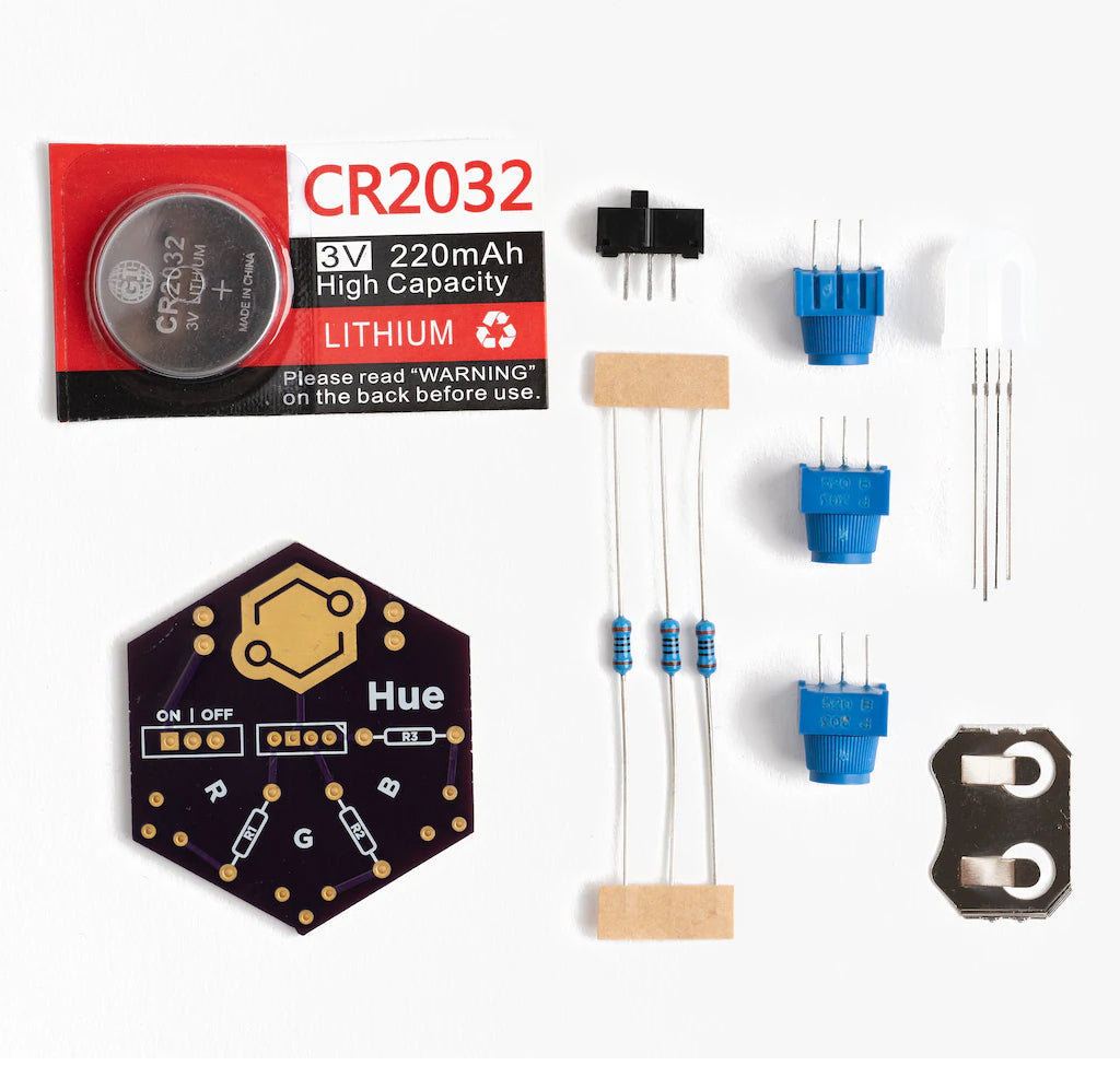 All of the parts for the kit are laid out; there is a silver cell battery in red and white packaging with a transparent window, one black and gold hexagon-shaped hue board, three blue and silver resistors, three light blue potentiometers, and one white RGB LED and a silver battery holder.