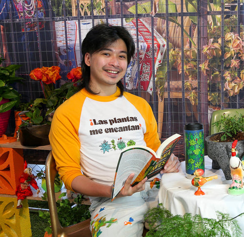 Person seated wearing a baseball style t-shirt with gold sleeves  that has succulents and ¡Las plantas me encantan! printed on the front.