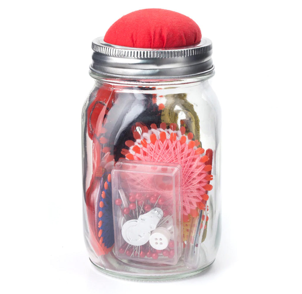A clear mason jar with a silver lid and a red pin cusion on the top with a variety of thread wheels, a thimble, stitch ripper, needle set, scissors, pins and buttons, needle threader, and a tape ruler. Unscrew the pin cushion lid to reveal an entire sewing kit. Unscrew the pin cushion lid to reveal an entire sewing kit.