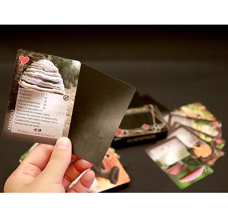 A stack of cards is fanned out on the table; someone holds up two cards, one with a mushroom and identifying facts, and a blank black card.