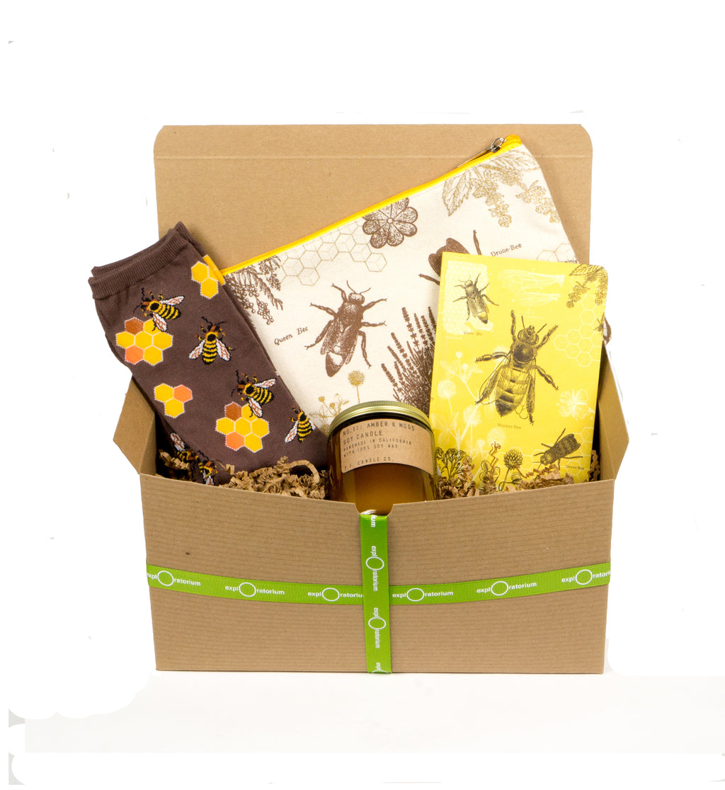 A natural colored box with green ribbon sits open with brown & yellow bee socks, a yellow bee-inspired journal, an amber glass candle, and a natural colored canvas pouch screen printed in brown with bees, honeycomb, and flowers.