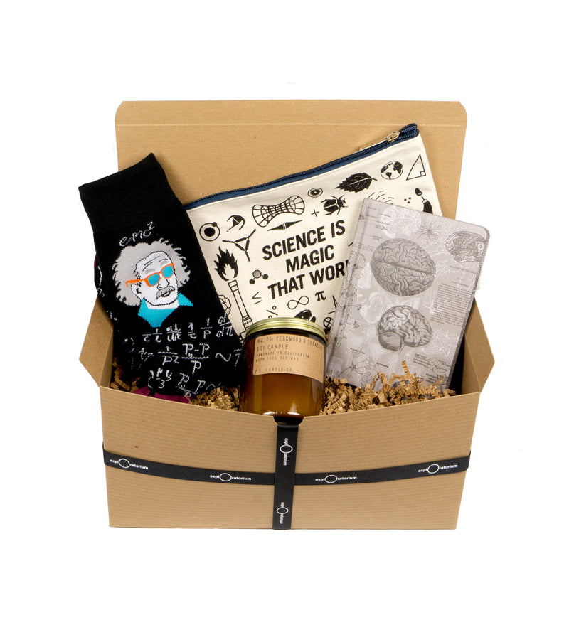 A natural colored box with a black ribbon sits open with black Einstein portrait socks, a grey brain-inspired journal, an amber glass candle, and a natural colored canvas pouch screen printed in black with Science is Magic and science tool icons.