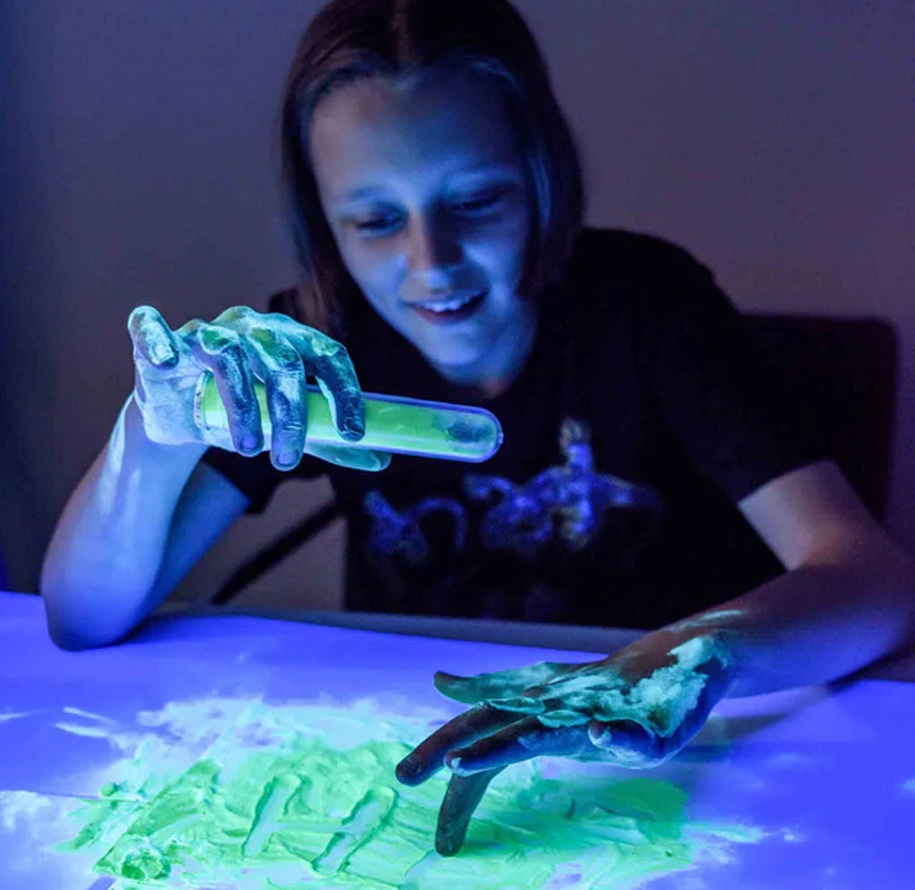 A girl is sitting in a dark room with glowing green phosphorescent powder on the table and her arms. A blue light shines, emphasizing the luminous aspects of the powder.