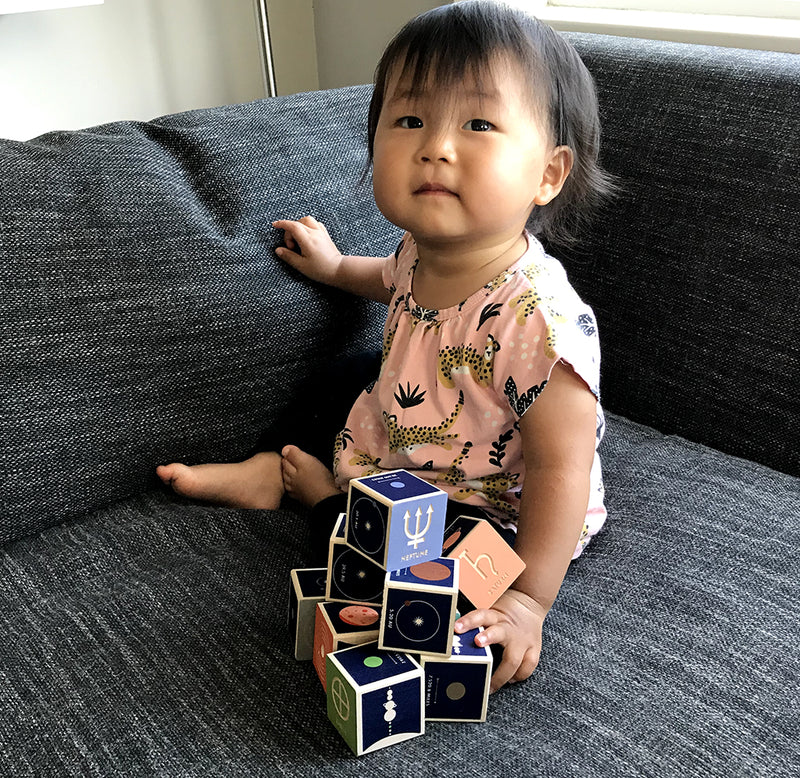 A young child sits on a couch looking toward the camera; the dark blue blocks with planets are stacked in front of her. 