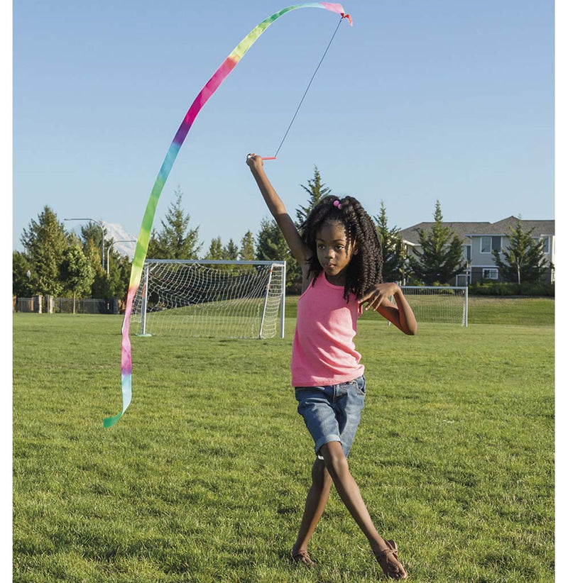 A young girl is standing in a park setting, twirling the rainbow-colored stunt streamer above her head.