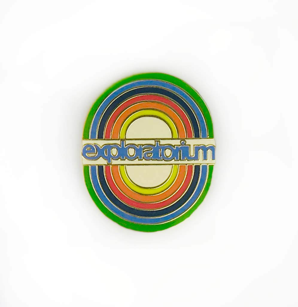Oval pin with rainbow circles. The word "exploratorium" in blue on a white rectangle is in the middle of the pin.