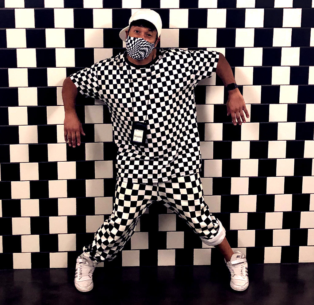 A man wearing a black and white checked shirt and pants and a black and white face mask is leaning back against a black and white checked wall.
