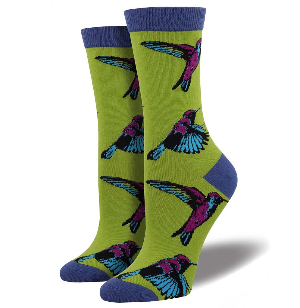 The socks are green with purple on the heel, toe, and top edge. There are four blue and magenta hummingbirds in different stages of flight moving up the socks.