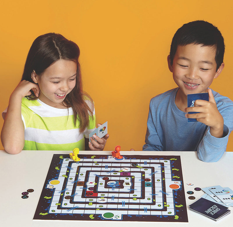 Two children are smiling with cards in their hands; the gameboard sits in front of them, set up for play. 