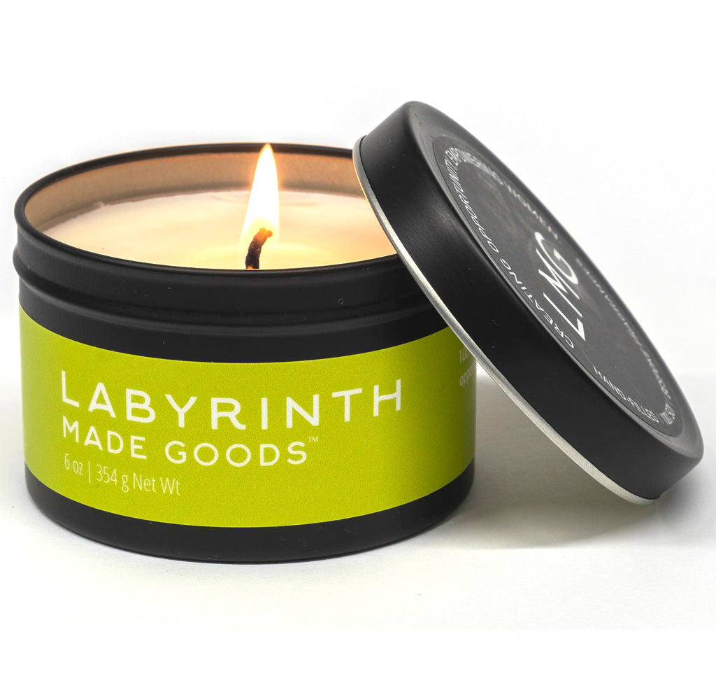 The tin for the candle is black with a green label with white text that reads Labyrinth Made Goods; the white candle is lit.