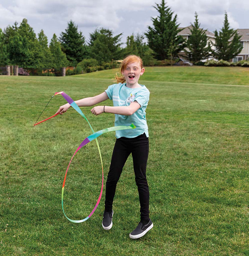 A young girl is standing in a park setting, looping the rainbow-colored stunt streamer in front of her.