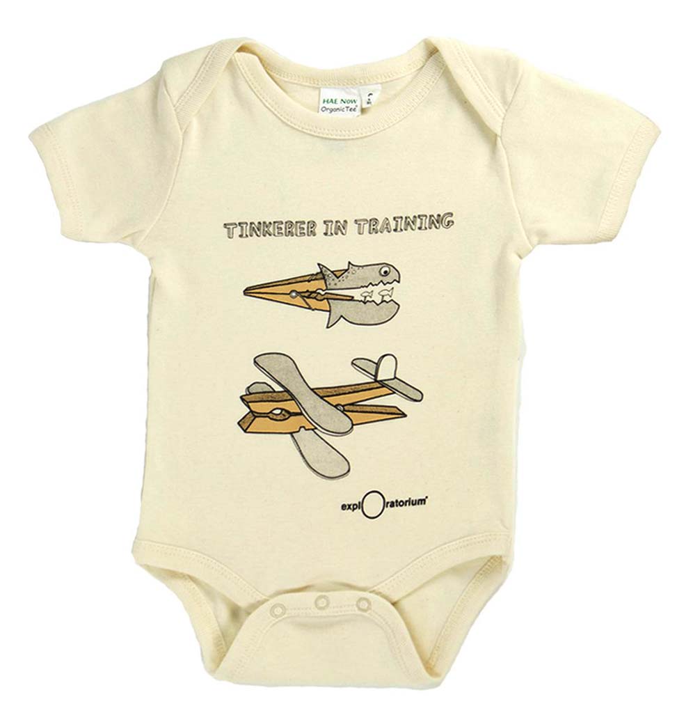 A cream-colored baby onesie. On the front, it reads "Tinkerer in Training." Under that are a grey paper fish attached to a close pin, a grey paper plane connected to a close pin, and the Exploratorium in black ink.