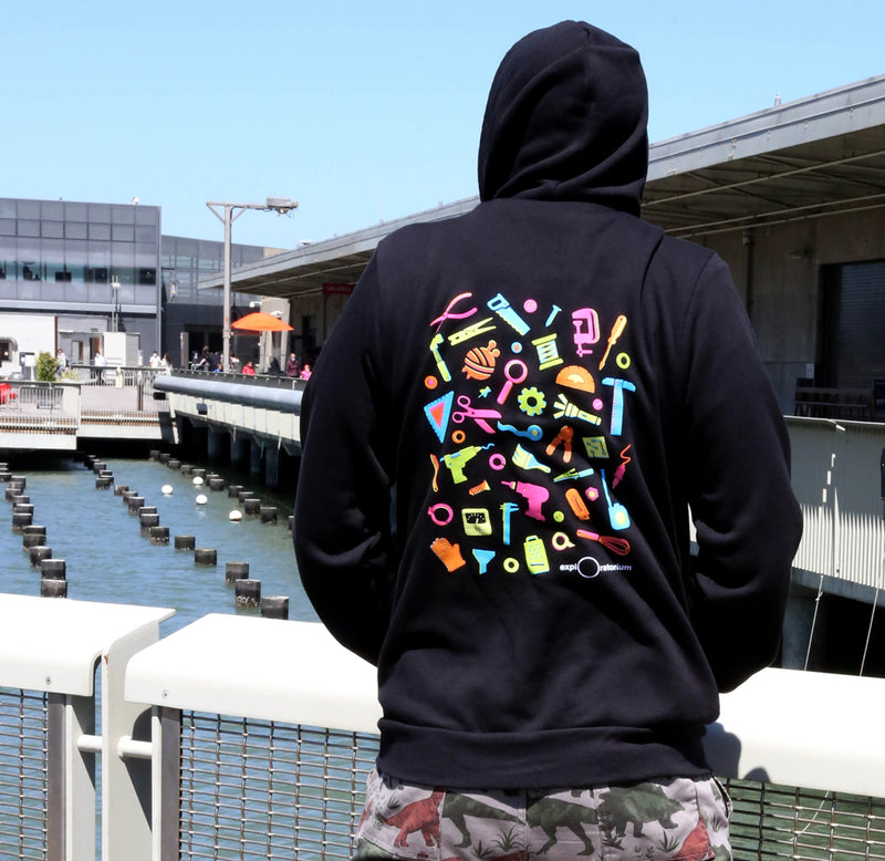 A person is standing with his back facing us and wearing the black Art of Tinkering hoodie with the neon tools blazoned on the back in colors of yellow, blue, pink, green, and orange.