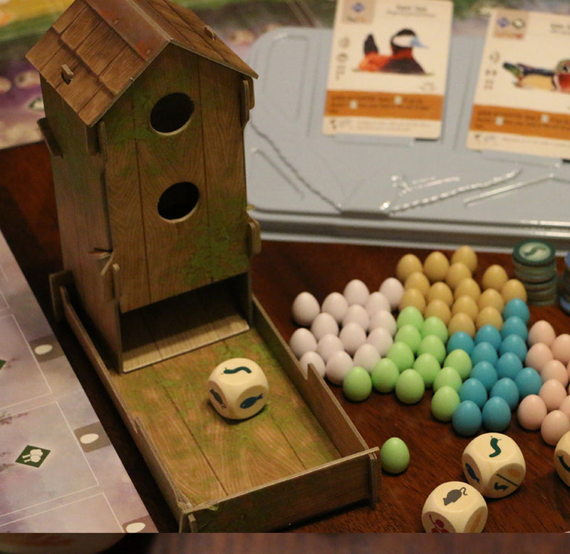 A wooden birdhouse sits in a wood box. One six-sided die sitting in the box game cards with birds, white eggshell blue, yellow and white eggs, game tokens, and gameboard sit on the table.