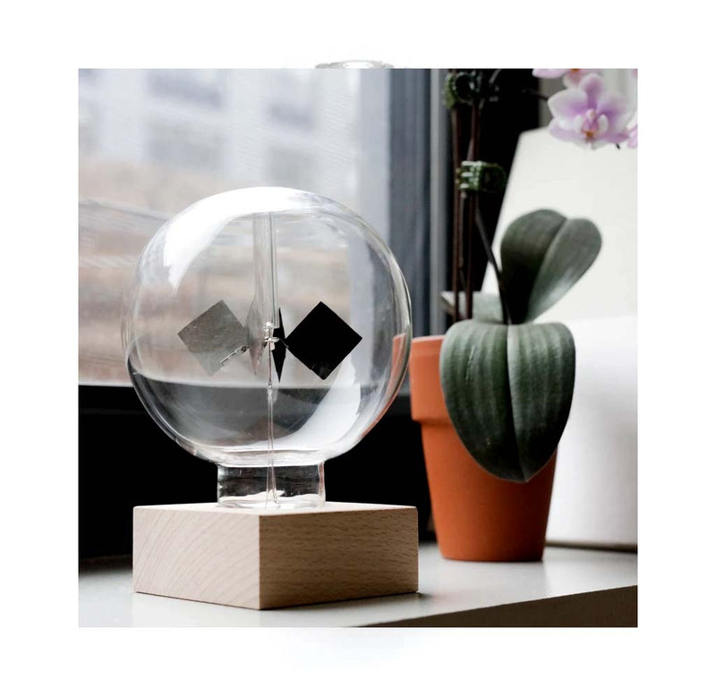 A clear circular globe sits atop a wooden base with four black triangular-shaped discs that rotate in the middle sitting on a desk next to a window,